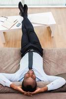 relaxamento total. vista superior do jovem africano feliz em trajes formais, deitado no sofá com as pernas em uma mesa e as mãos atrás da cabeça foto