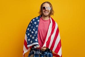 jovem bonito coberto com a bandeira americana, olhando para a câmera e sorrindo contra um fundo amarelo foto