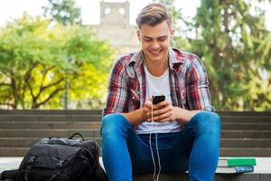 curtindo sua música favorita. feliz estudante do sexo masculino ouvindo mp3 player e sorrindo enquanto está sentado na escada ao ar livre com livros e mochila perto dele foto