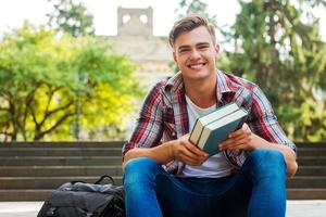 feliz leitor de livros. estudante do sexo masculino bonito segurando livros e sorrindo enquanto está sentado na escada ao ar livre com o prédio da universidade ao fundo