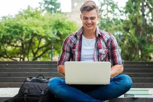 navegar na net ao ar livre. feliz estudante do sexo masculino trabalhando no laptop e sorrindo enquanto está sentado na escada ao ar livre com livros e mochila perto dele foto