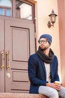 desfrutando de bebida quente. jovem bonito com roupa casual inteligente, sentado em frente à entrada da casa e segurando a xícara de café foto