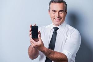 apresentando novo smartphone. homem maduro confiante de camisa e gravata mostrando seu novo celular e sorrindo em pé contra um fundo cinza foto