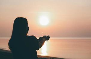 silhueta de mãos rezando com deus de frente para o céu ao nascer do sol na praia, fé na religião e crença em deus, poder da esperança ou amor e devoção. foto