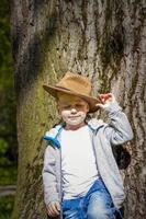 menino bonito posando com um chapéu de cowboy na floresta por uma árvore. os raios do sol envolvem o espaço. história de interação para o livro. espaço para copiar foto