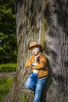 menino bonito posando com um chapéu de cowboy na floresta por uma árvore. os raios do sol envolvem o espaço. história de interação para o livro. espaço para copiar foto