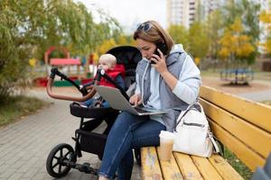 uma jovem mãe sentada em um banco ao lado de um carrinho com uma criança trabalha on-line em um laptop e atende um telefonema foto