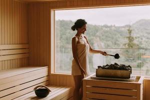 jovem mulher despejando água em pedra quente na sauna foto