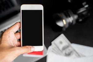 mão esquerda segurando smartphone em borrão blackground com laptop, dólares, câmera e cartão de crédito foto