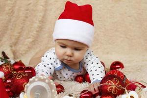 menina bonitinha no chapéu vermelho de papai noel está brincando com decorações de natal brilhantes vermelhas e brancas e luzes de natal em uma manta bege. foto