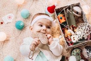 menina bonitinha de vestido azul e branco está brincando com coração de madeira em uma manta bege com luzes e decorações de natal, vista superior. foto