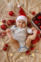 adorável menina sorridente no chapéu vermelho de papai noel está deitada em uma manta bege com decorações de natal vermelhas e brancas e luzes de natal e segurando os dedos dos pés nas mãos, vista superior. foto