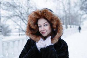 retrato de uma jovem mulher bonita com olhos azuis com casaco de pele preta com capuz vermelho em uma caminhada no parque de inverno nevado. foto