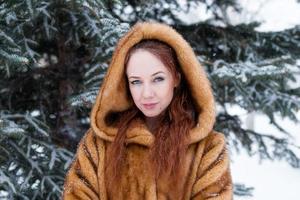 retrato de uma jovem mulher bonita com cabelo vermelho e olhos azuis com casaco de pele falsa no fundo do parque de inverno nevado. foto