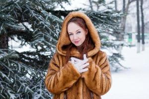 jovem mulher bonita com cabelo vermelho e olhos azuis com casaco de pele falsa está andando no parque de inverno nevado. foto