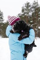 jovem está segurando seu schnauzer miniatura preto congelado pelas mãos em um fundo de floresta de coníferas de inverno. foto