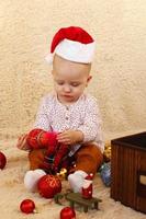 menina de chapéu vermelho de papai noel está brincando com decorações de natal em uma manta bege. foto