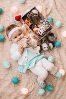 linda garotinha sorridente de vestido azul e branco está brincando com coração de madeira em uma manta bege com enfeites de natal e luzes, vista superior. foto