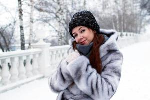 jovem mulher bonita com cabelo vermelho e olhos azuis com casaco de pele cinza com capuz está andando no parque de inverno nevado. foto