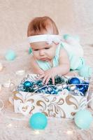 menina bonitinha de vestido azul e branco está brincando com decorações de natal de uma caixa em uma manta de pelúcia bege com luzes de natal. foto