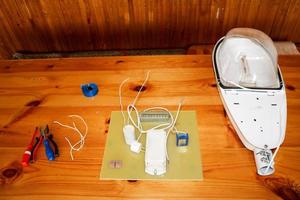 uma grande lâmpada de rua branca desmontada com um circuito elétrico com fios e peças de reposição, equipamento de instalação, alicate, fita isolante azul, chaves de fenda na mesa foto