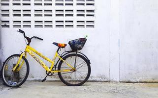 bangkok, tailândia-25 de dezembro de 2020 bicicleta amarela velha estacionada contra a parede de concreto com garrafa de plástico ou despejo de resíduos na cesta preta na traseira e pneu furado com espaço de cópia. bicicleta vintage ou quebrada. foto