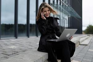 uma mulher de negócios de meia-idade trabalha online em um laptop contra o pano de fundo de um prédio de escritórios foto