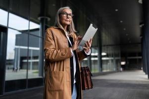 elegante empresária idosa com uma maleta e um tablet nas mãos na entrada do centro de negócios foto