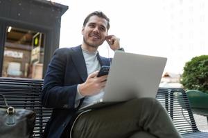 um homem com um sorriso largo no rosto ouve música em fones de ouvido sentado por telefone e laptop ao lado de um café, reunião on-line de conceito foto