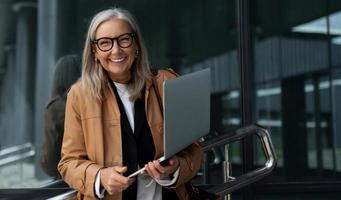 empresária idosa satisfeita com um sorriso largo e um laptop nas mãos contra o pano de fundo do centro de escritórios foto