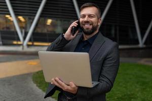 homem muito adulto em um terno de negócios com um laptop nas mãos fala em um telefone celular no fundo do centro de escritórios, conceito de um corretor de imóveis de sucesso foto