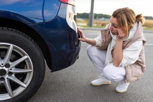 motorista feminina frustrada senta-se no pára-choque traseiro do carro e examina os danos e arranhões, conceito de acidente de trânsito foto