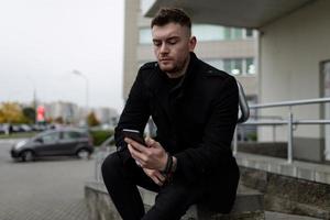 jovem brutal em um casaco preto com um telefone celular no fundo do centro de escritórios foto