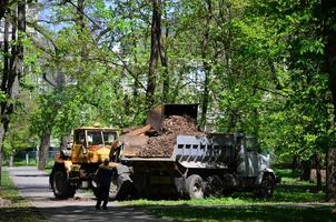 a equipe de melhoria da cidade remove as folhas caídas no parque com uma escavadeira e um caminhão. trabalho sazonal regular na melhoria dos locais públicos para recreação foto