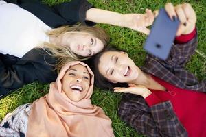 namoradas multirraciais rindo enquanto estava deitado no gramado e tomando selfie foto