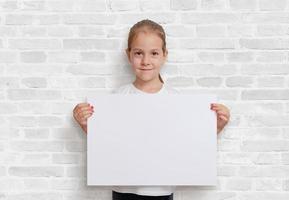 garota segurando um cartaz na posição horizontal. papel em branco para apresentação do projeto. parede de tijolos brancos no fundo foto