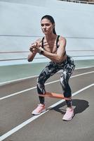 sem ifs ou buts para treino. mulher jovem e bonita em roupas esportivas agachada usando banda de resistência em pé na pista de corrida ao ar livre foto