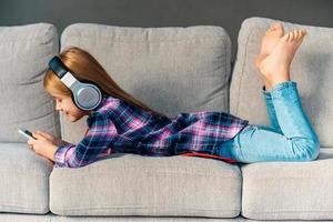 pequeno amante da música. linda garotinha em fones de ouvido segurando seu telefone inteligente com sorriso enquanto estava deitado na frente no sofá em casa foto