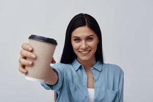 mulher jovem e atraente segurando o copo descartável e sorrindo em pé contra um fundo cinza foto