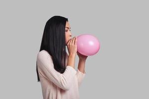 em um clima para um grande feriado. tiro de estúdio de mulher jovem e atraente em roupas casuais, explodindo um balão rosa em pé contra um fundo cinza foto