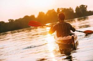 apreciando o pôr do sol no rio. vista traseira do jovem andando de caiaque no lago com pôr do sol ao fundo foto