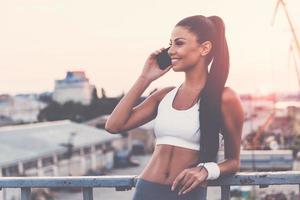 compartilhando suas conquistas com o amigo. mulher jovem e bonita em roupas esportivas falando no celular e sorrindo em pé na ponte com a luz do sol da noite e vista urbana ao fundo foto