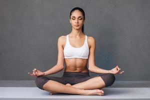 longa respiração. bela jovem africana em roupas esportivas praticando ioga enquanto está sentado em posição de lótus contra fundo cinza foto