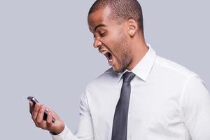 notícia muito ruim. furioso jovem africano de camisa e gravata segurando o celular e gritando em pé contra um fundo cinza foto