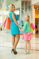 mãe e filha fazendo compras. alegre cabelo loiro mãe e filha segurando sacolas de compras e olhando por cima do ombro em pé no shopping foto
