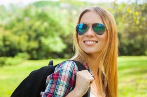 belo caminhante. mulher jovem e bonita em óculos de sol carregando sua mochila e olhando por cima do ombro em pé na natureza foto