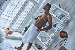 manhã fresca. vista superior do jovem africano bonito coberto com toalha de banho segurando uma xícara e mantendo a mão no quadril em pé no quarto em casa foto