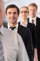 confiante e bem sucedido. jovem confiante em trajes formais olhando para a câmera e sorrindo enquanto está na fila com outras pessoas foto