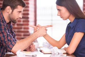 culpando um ao outro. imagem de vista lateral de homem bravo e mulher sentada cara a cara na mesa do escritório e apontando um ao outro foto