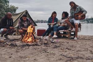 tocando música favorita. grupo de jovens em roupas casuais sorrindo enquanto desfruta de festa na praia perto da fogueira foto